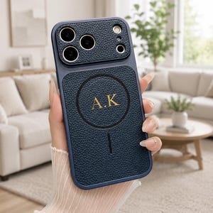 Puede incluir: Una funda de teléfono azul marino con acabado de piel sintética texturizada y borde liso. La parte trasera presenta un anillo estilo MagSafe con las iniciales A.K grabadas en oro en el centro y una línea vertical en la parte inferior.