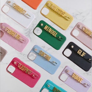 Puede incluir: Surtido de fundas de teléfono de colores con nombres personalizados y un símbolo de corazón en letras doradas. Cada funda tiene una correa con un anillo dorado.