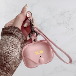 May include: A pink leather case for AirPods with a black strap and a gold clasp. The case has the initials "MM" embossed on the front.