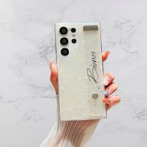 May include: A pearlescent white phone case with a rectangular design. The case features a camera array, the word "Piones" in script, and the word "fashion" in a small rectangular accent. A silver button is also visible.