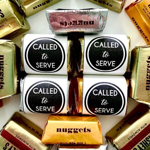 Puede incluir: Primer plano de varias pequeñas barras de chocolate envueltas en papel de aluminio dorado. Las barras están etiquetadas como "nuggets" y "Hershey's". También hay varias etiquetas blancas con texto negro que dicen "Called to Serve".
