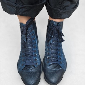 May include: A pair of high-top lace-up boots in shades of dark blue and black. The boots feature a textured leather finish, a rounded toe cap with subtle perforated detailing, and matching dark blue laces threaded through metal eyelets.