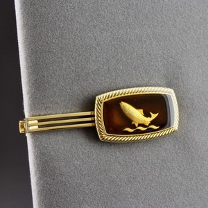 May include: Gold tie clip with a rectangular amber-coloured stone featuring a gold fish jumping out of the water.