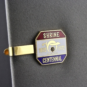 May include: A gold tie bar with a red, white, and blue enamel emblem. The emblem says "Shrine Centennial 1872-1972 AAONMS".