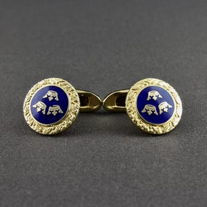 May include: A pair of gold cufflinks with a blue enamel design featuring three stylized crowns. The cufflinks have a textured gold border.