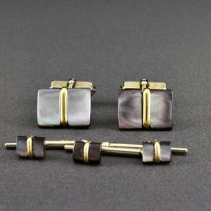 May include: A set of two rectangular cufflinks and three matching shirt studs, each featuring iridescent dark gray mother-of-pearl inlays divided by a central vertical gold-tone metal bar, showcasing a polished finish and a classic rectangular shape.