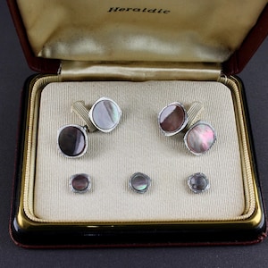 May include: A set of cufflinks and shirt studs presented in a brown box. The cufflinks and studs feature a silver-tone metal frame with a mother-of-pearl inlay. The box is lined with cream fabric and has the word "Heraldic" printed on the inside of the lid.