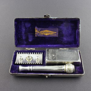 May include: A vintage Gillette razor set in a silver-colored case with a purple velvet lining. The set includes a razor head, a handle, and a blade dispenser. The case has a Gillette logo on the inside.