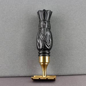 May include: A black and gold safety razor with a detailed handle. The handle features a carved figure of a man with a long beard and crown. The razor head and base are gold-colored.