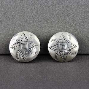 May include: A pair of silver earrings with a geometric design. The earrings are round and have a raised design of a square with four points.