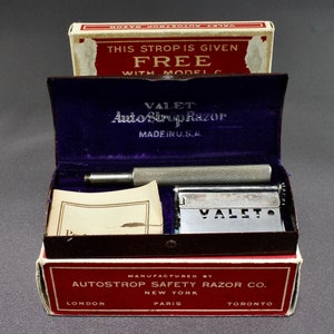 May include: A vintage Valet Auto Strop Razor set in its original box. The box is red and white with the words "Auto Strop Safety Razor Co." printed on it. The razor is silver and has the word "Valet" engraved on it. The set also includes a strop, which is a leather strap used to sharpen the razor blade.