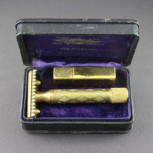 May include: A gold-colored Gillette New Standard safety razor and blade in a black leather case with a purple velvet lining. The case has the words "Gillette New Standard" printed on the inside.