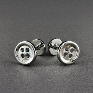 May include: A pair of silver button-shaped cufflinks. Each cufflink has a circular face with four small holes, resembling a button. The cufflinks are set against a dark gray background.