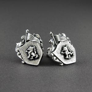 May include: A pair of silver-toned metal cufflinks featuring a shield-shaped design. Each cufflink displays a raised lion figure in the centre, topped with a small flag element and decorative scrollwork along the edges with a textured finish.