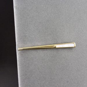 May include: A gold-toned tie bar featuring a slender, tapered design with a rectangular white mother-of-pearl inlay set into the wider end. The metal surface has a polished finish, and the clip is positioned horizontally.
