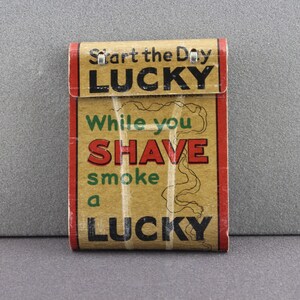 May include: A vintage Lucky Strike cigarette pack with a brown background and red and green accents. The text on the pack reads "Start the Day Lucky" and "While you Shave smoke a Lucky".