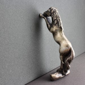 May include: A silver-toned bottle stopper in the shape of a rearing horse. The horse is detailed with a flowing mane and tail, and is posed with its front legs raised. The stopper is designed to fit into a bottle.
