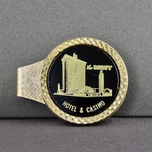 May include: A gold-coloured metal money clip with a black circular centre. The centre features a gold illustration of a building with the text "The MINT HOTEL & CASINO". The clip has a textured design.
