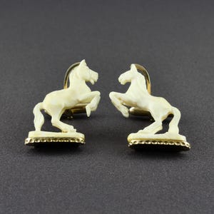May include: A pair of cufflinks featuring off-white, carved horse figures in a rearing position. Each horse is mounted on a gold-toned base with a textured edge, and the metal hardware is visible behind the horse figures on the back side.