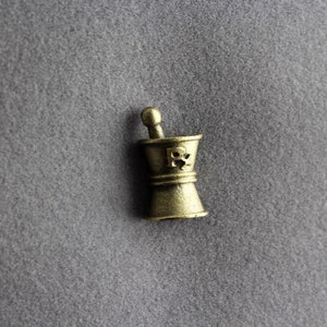 May include: A gold-colored metal mortar and pestle with a skull and crossbones design. The mortar and pestle are a traditional tool used for grinding herbs and spices.