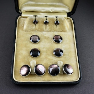 May include: A set of black and iridescent mother-of-pearl cufflinks and shirt studs displayed in a black velvet-lined box. The set includes various sizes of round buttons, showcasing a range of colors from dark grey to purple.