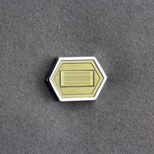 May include: A silver and gold hexagonal pin with a rectangular design in the center. The pin has a textured surface and a subtle sheen.