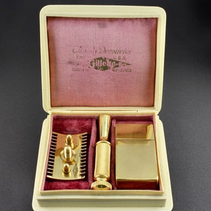 May include: A vintage Gillette razor set in a cream-colored box with a pink velvet interior. The set includes a gold-plated razor, a gold-plated handle, and a gold-plated blade holder. The box is labeled "Safety Razor Gillette" and "Made in U.S.A."
