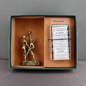 May include: A bronze-colored sculpture of two dancers in a green-edged box with a brown interior. The box also contains a clear plastic package with text that reads "DANCE RAZOR" and "C.A.DANIELS & Company".
