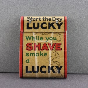 May include: Vintage Lucky Strike cigarette pack advertising with the text "Start the Day Lucky, While You Shave Smoke a Lucky". The pack is brown with red and green accents.