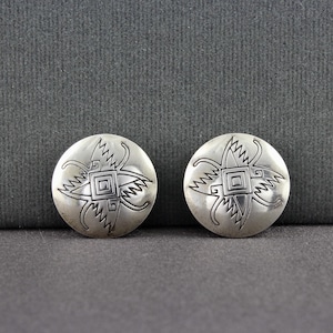 May include: Two silver-toned stud earrings. Each earring is round with a detailed, symmetrical design featuring a central square and radiating lines. The earrings are displayed on a dark gray surface.
