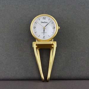 May include: A gold-tone Neiman Marcus quartz watch with a round white face, black numerals at 3, 6, and 12, and black hands. The watch is attached to a gold-tone metal clip with a triangular, open-frame design, suitable for attaching to clothing.