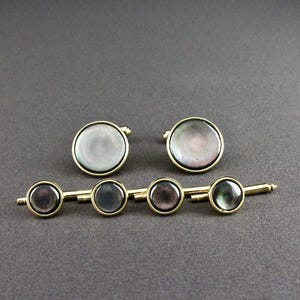 May include: A set of two round cufflinks and a matching four-button shirt stud set, each featuring a circular iridescent mother-of-pearl inlay framed in a polished gold-tone metal setting, designed for formal attire and shirt closures.