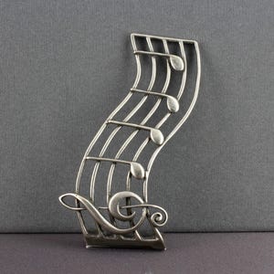 May include: A silver-toned metal musical note brooch. The brooch features a stylized treble clef and musical notes on a curved staff. The design is detailed and has a vintage aesthetic, perfect for music lovers.