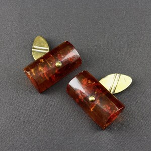May include: A pair of vintage cufflinks with rectangular, reddish-brown marbled faces and gold-toned metal accents. Each cufflink features a small, round gold-toned stud and a gold-toned, leaf-shaped backing.