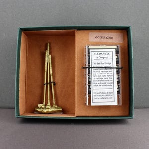 May include: A gold-colored golf club-themed razor set in a green and brown box. The set includes a miniature golf club sculpture and a package of razor cartridges. The text "GOLF RAZOR" is visible on the box.