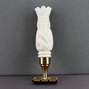 May include: A white, decorative razor with a detailed handle featuring a carved figure wearing a crown. The handle is connected to a gold-colored metal base and blade. The razor is set against a gray background.