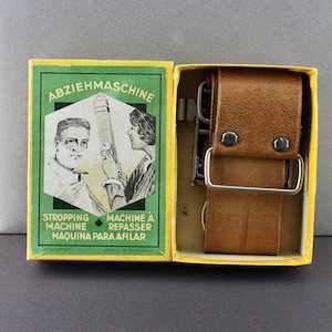 May include: A green and yellow box with the text "Abziehmaschine" and illustrations of a man and woman using a razor strop. The box contains a brown leather razor strop with a metal buckle.