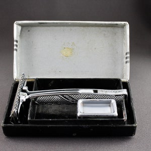 May include: A silver safety razor with a black plastic case. The razor has a chrome finish and a textured handle. The case has a black velvet lining.