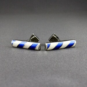 May include: A pair of bar-shaped cufflinks featuring a diagonal striped pattern in alternating royal blue and cream enamel. The metal frame is polished silver-tone, and each cufflink includes a hinged T-bar toggle mechanism on the back for fastening.