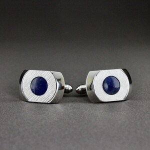 May include: A pair of silver-toned metal cufflinks featuring a rectangular shape with rounded sides and a brushed finish. Each cufflink is centered with a smooth, circular, deep blue stone inlay, providing a contrast to the metallic surface.