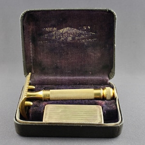 May include: A gold-plated safety razor in an open black leather case. The razor has a gold-plated handle and a gold-plated head with a safety bar. The case is lined with black velvet and has a gold-plated inscription that reads "MADE IN U.S.A.  THE GEM DAMASCUS SAFETY RAZOR CORP.  NEW YORK, N.Y."