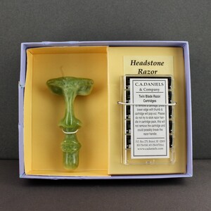 May include: A green, headstone-shaped razor handle with a matching box. The box contains a clear plastic case with twin blade razor cartridges and the text "Headstone Razor" and "C.A.DANIELS & Company".