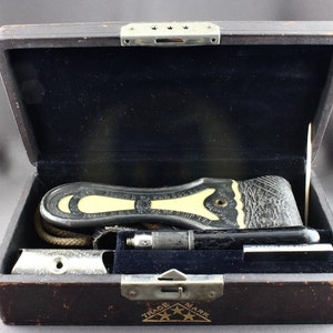 May include: A black leather case with a silver latch and a gold inscription that reads "TRADE MARK" with three stars. The case is open and contains a black and white leather razor with a yellow handle, a silver razor blade holder, a black and silver razor handle, and a black and silver razor blade.