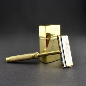 May include: A gold-colored safety razor with a gold-colored case. The razor has a white plastic blade and the case has the text "The Gillette " printed on it.