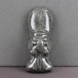 May include: A carved stone hippo figurine in shades of gray and black. The hippo is standing upright with its head and feet clearly defined. The sculpture has a smooth, polished finish and is set against a gray background.