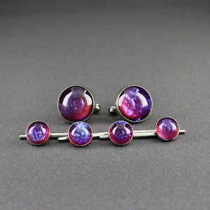 May include: A set of two round cufflinks and a tie bar featuring a galaxy-inspired design with swirling shades of deep purple, magenta, and blue. Each piece has a smooth, domed glass cabochon set in a polished silver-toned metal frame.