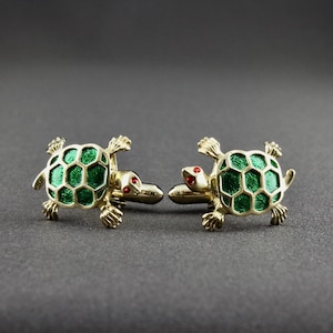 May include: A pair of gold-toned cufflinks shaped like turtles. The turtle shells are green with a honeycomb pattern, and the eyes are red. The cufflinks are set against a dark gray background.