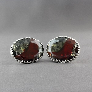 Op de afbeelding: Een paar ovale manchetknopen met een facetgeslepen, roodbruine edelsteen in het midden. De edelstenen zijn gezet in een zilverkleurig metaal met een decoratieve, geschulpte rand. Op een grijze achtergrond.