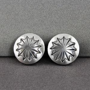 May include: Two silver-colored, round stud earrings. Each earring features a starburst design with etched lines radiating from the center. The earrings are set against a gray background, highlighting their metallic sheen and intricate details.
