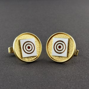 May include: A pair of gold cufflinks with a white square and red and white concentric circles in the centre.